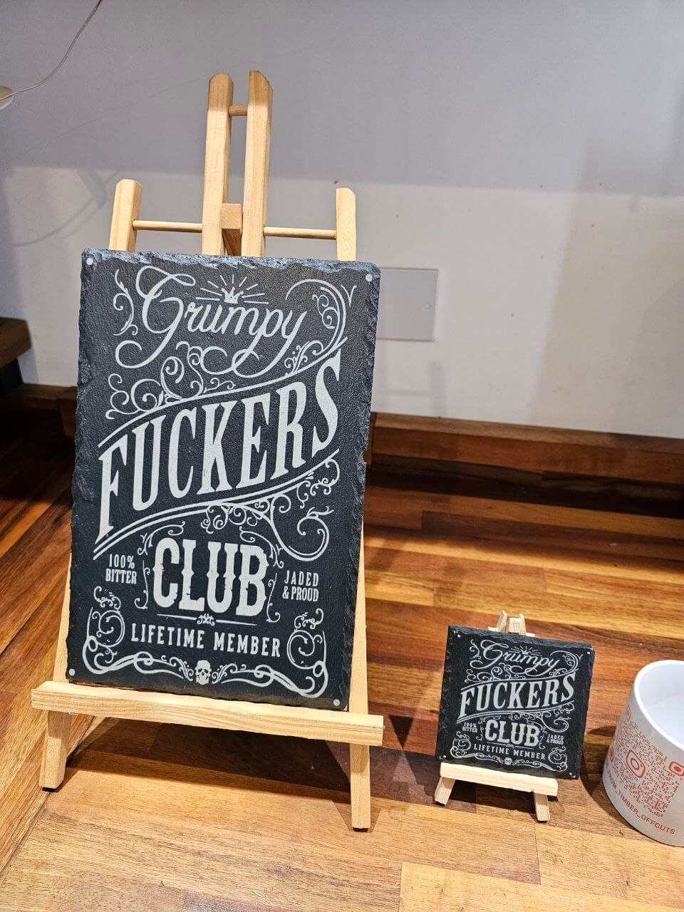 Slate coaster with 'Grumpy Fuckers Club' design displayed on an easel next to a coffee mug.
