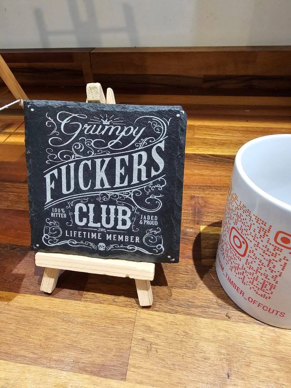 Slate coaster with 'Grumpy Fuckers Club' design displayed on an easel next to a coffee mug.